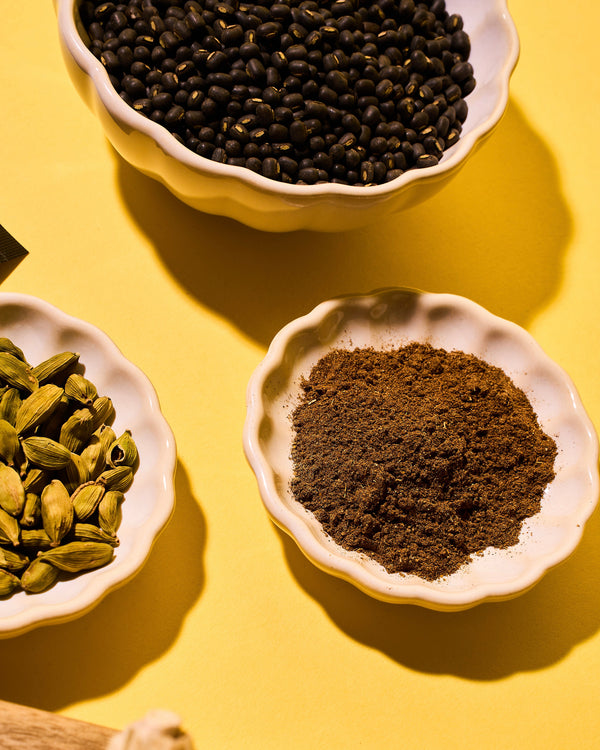 Three bowls containing black beans, cardamom pods, and ground coffee on a yellow background
